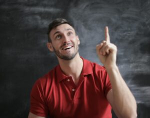 Un jeune homme heureux vêtu d'un polo rouge, souriant et pointant vers le haut devant un tableau noir.