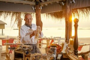 Chef préparant des pâtes fraîches au bord d'un coucher de soleil sur la plage, capturant l'art culinaire dans un cadre tropical.